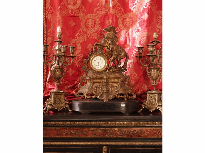 Trittico in antimonio dorato, orologio da tavolo sormontato da scena equestre con coppia di cande...