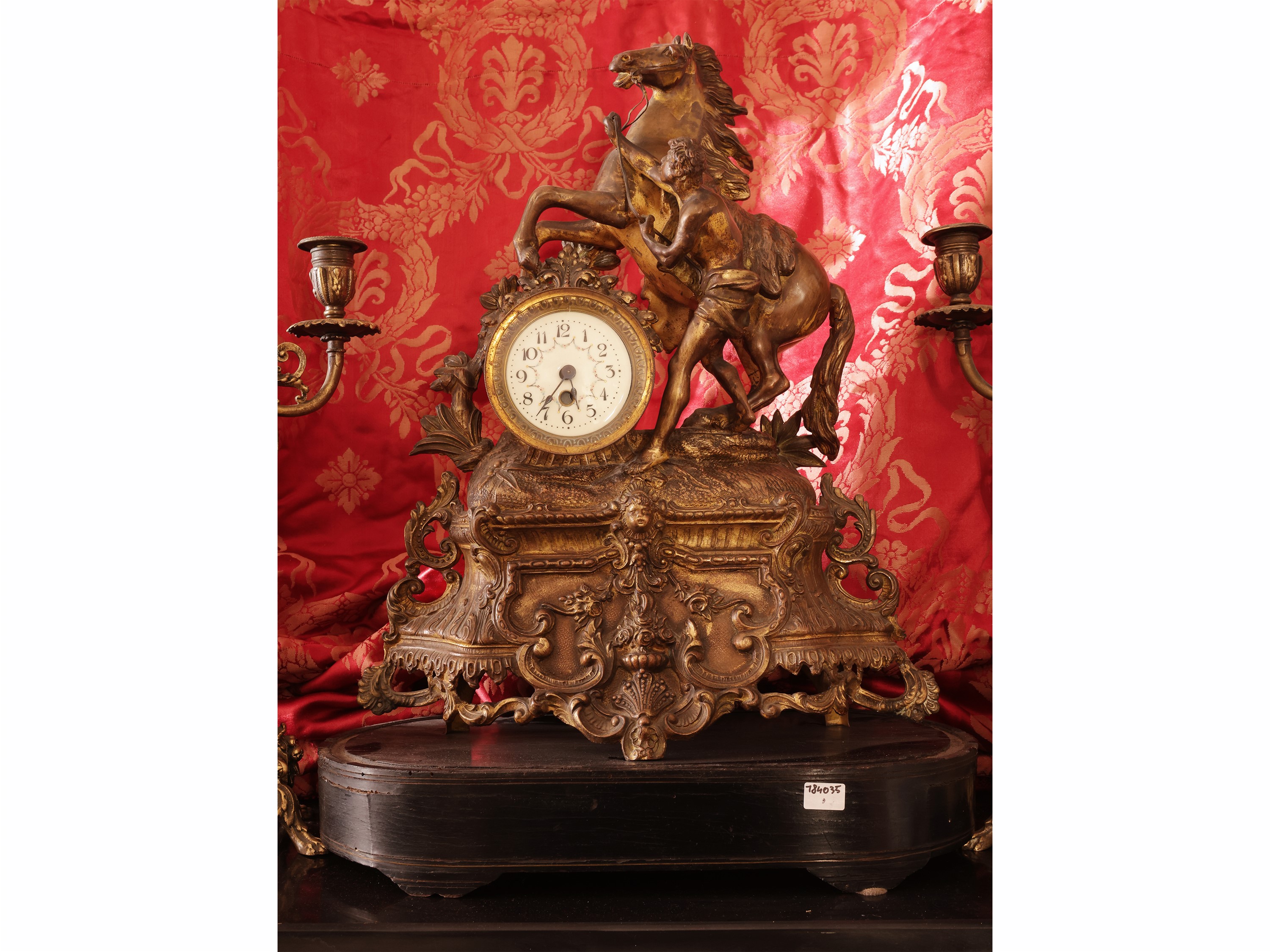 Trittico in antimonio dorato, orologio da tavolo sormontato da scena equestre con coppia di cande...